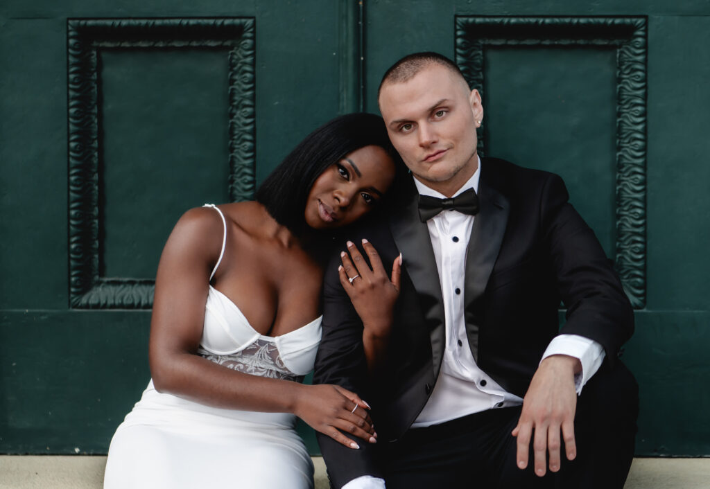 A stunning black bride in a white gown rests her head on her tux-wearing fiance's shoulder as they sit in front of a dark green door
