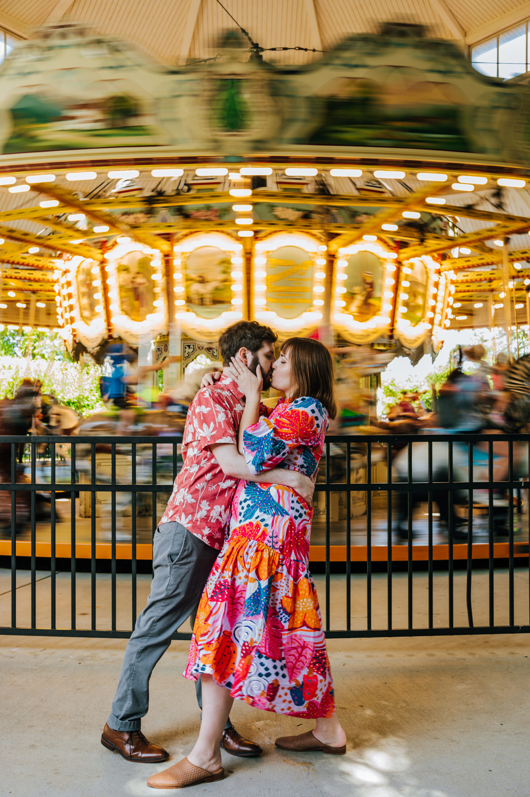 A couple wearing colorful clothing stops for a kiss in front of a spinning carousel