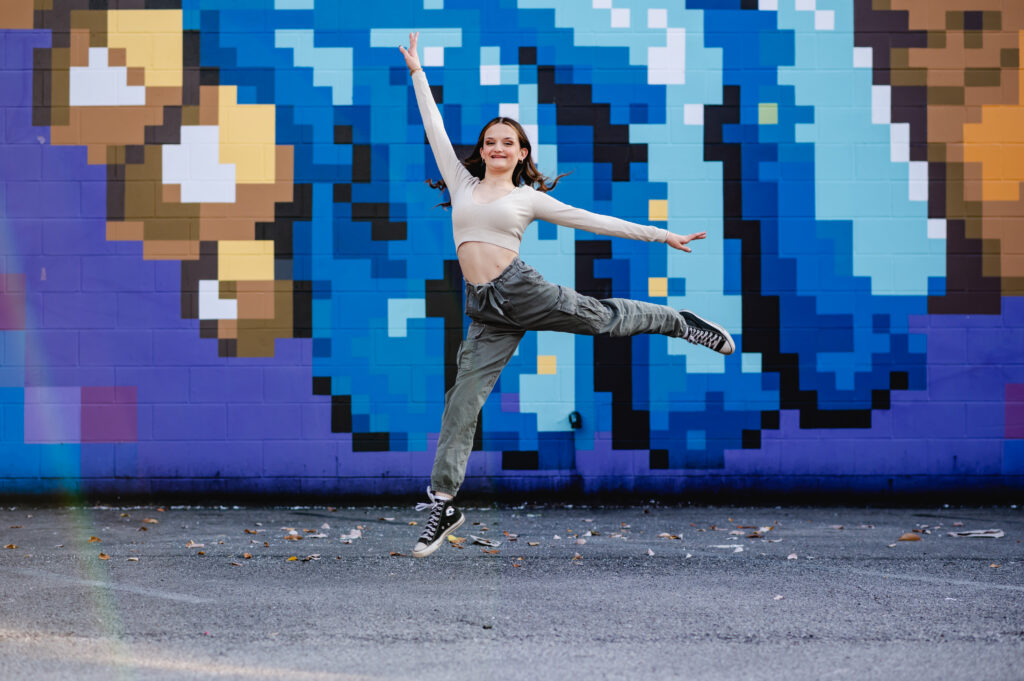 A high school senior girl leaps in the air in front of a pixel-pattern colorful mural