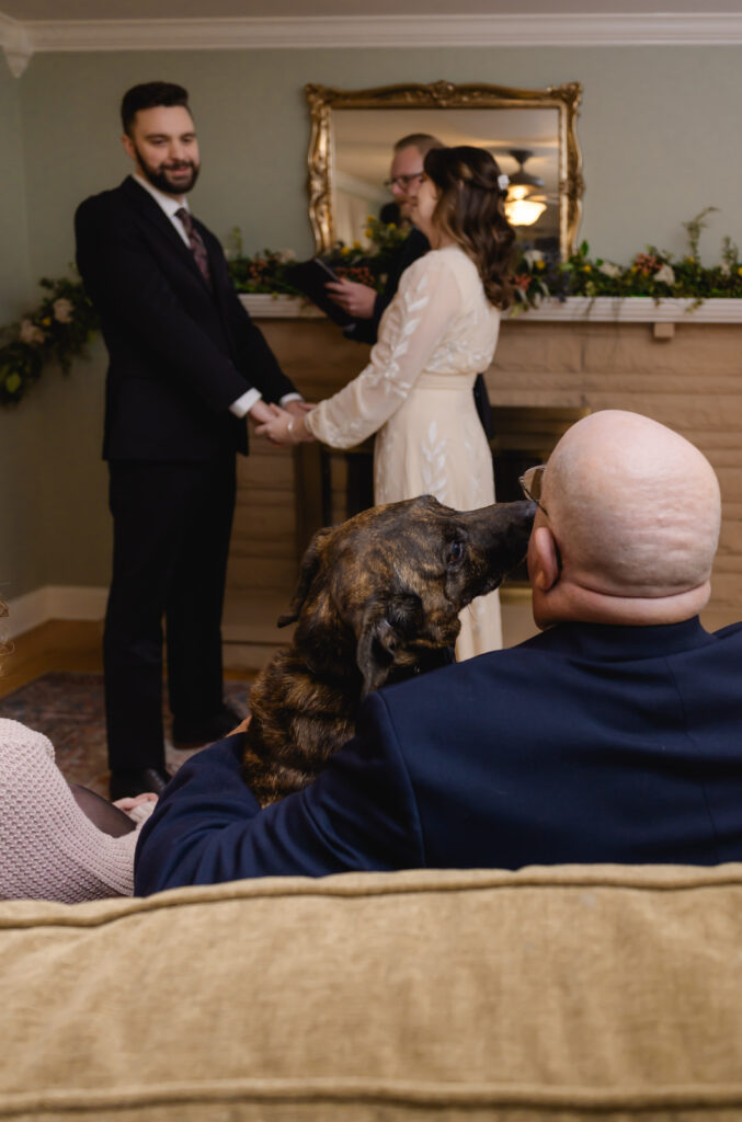 A bride's dad sits on the couch with her dog, who licks his face while the bride and groom get married in front of them