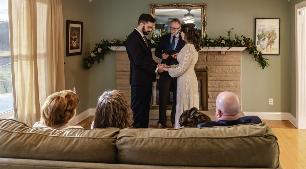 A couple and their wedding officiant stand in their living room for their home wedding while their family and dog watch from the couch