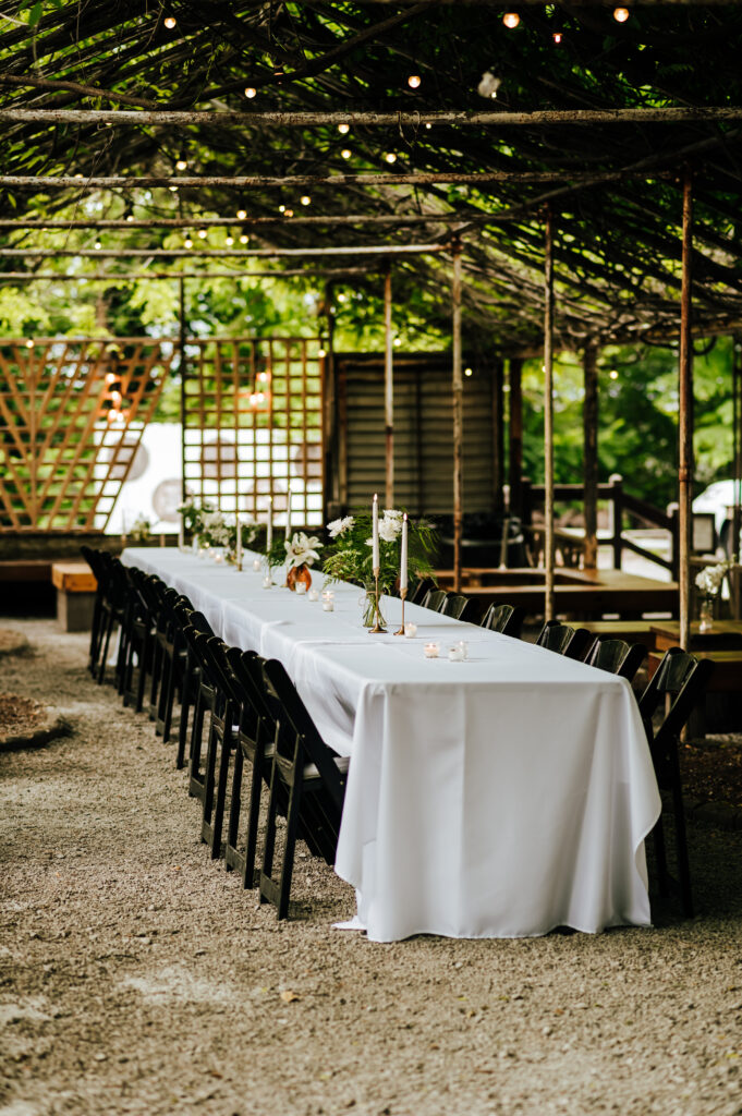 A long table with a white tablecloth, black chairs and candles sit underneath a wooden structure with string lights