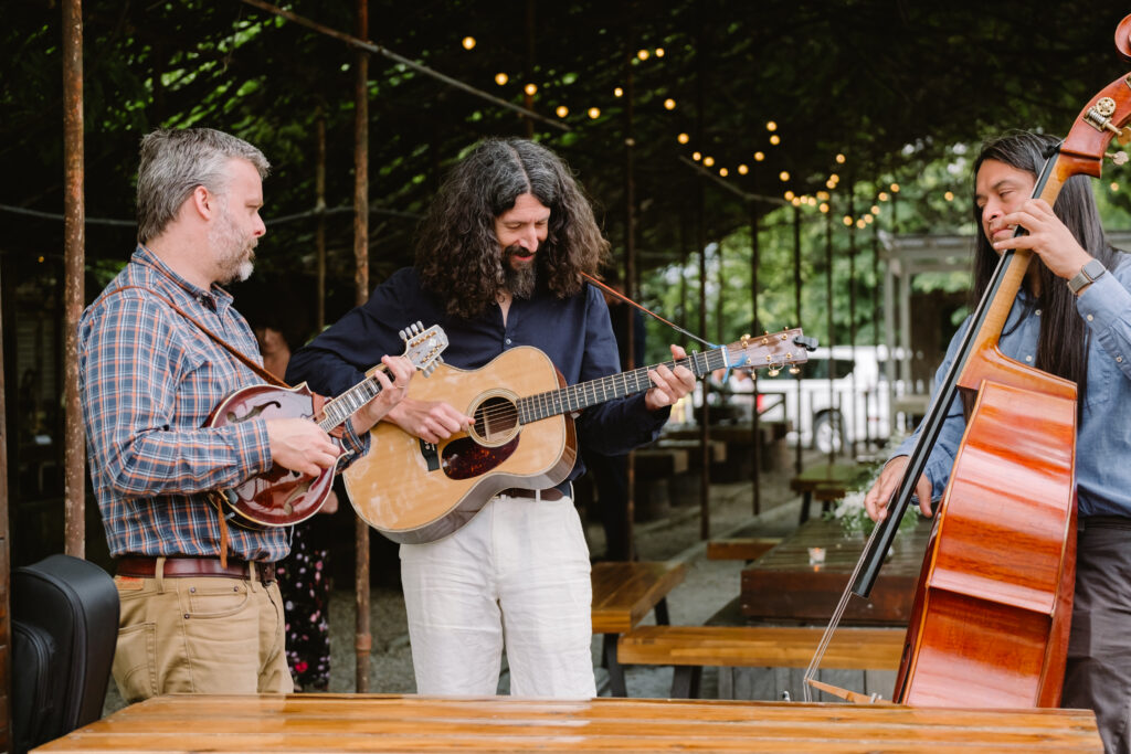 A three piece band plays guitar, banjo and cello at a cocktail hour outdoors under string lights