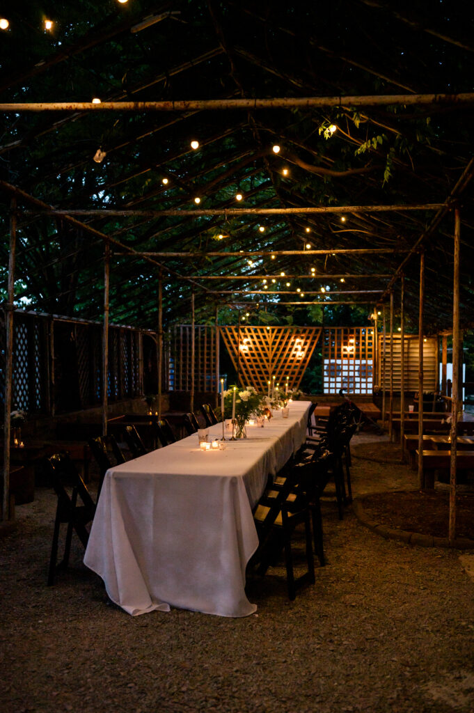 A long table with a white tablecloth, black chairs and candles sit underneath a wooden structure with string lights