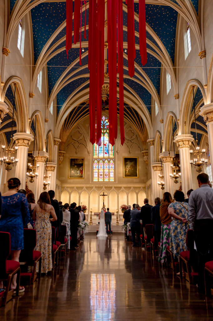 A wide angle photo shows the entire cathedral from floor to ceiling with the couple standing at the altar surrounded by their guests