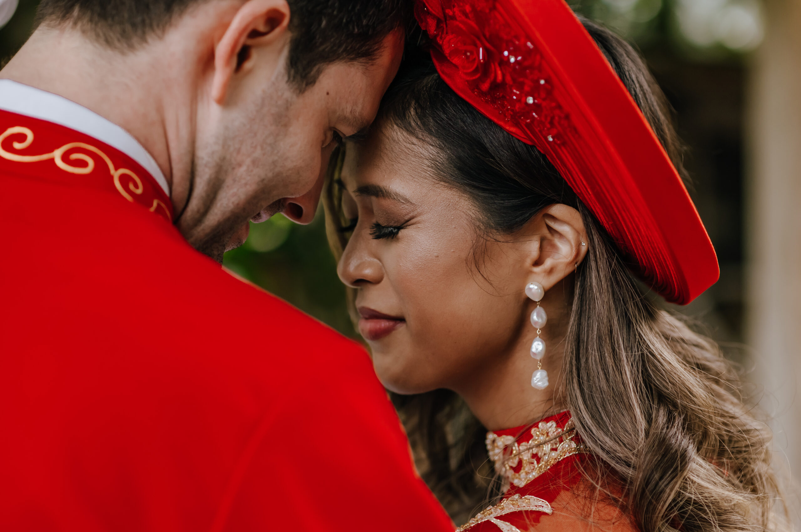 A couple rest their foreheads together with closed eyes while wearing traditional red ao gam and ao dai (Vietnamese wedding attire)