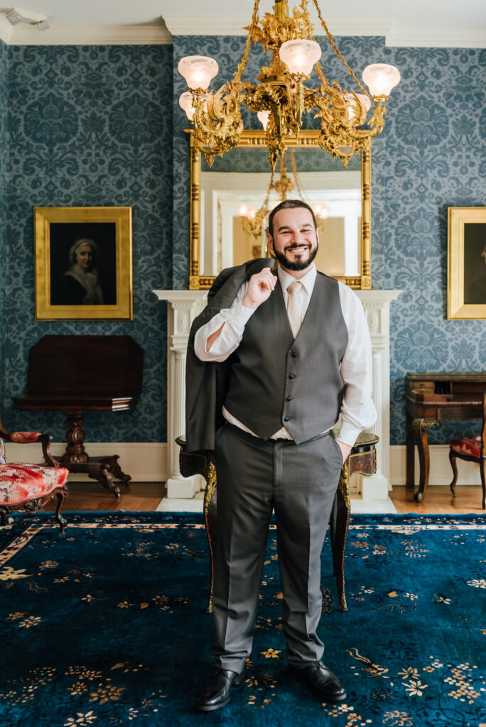 A groom poses for a portrait with his jacket over his shoulder in a stately blue room
