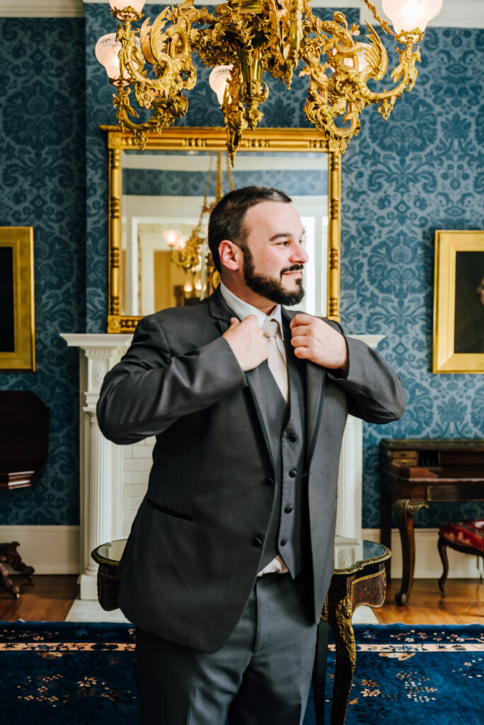 A groom adjusts his gray suit jacket in a blue and gold historic room