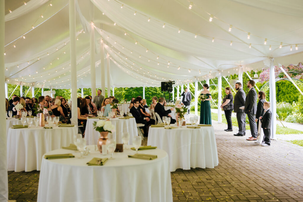 The groom and guests stand in a tented ceremony space waiting for the bride to enter.