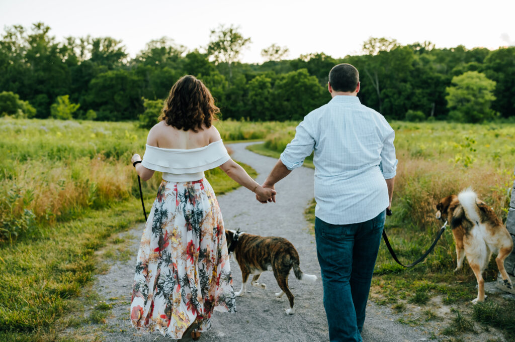 A couple holds hands and walks down a winding path with their two dogs.