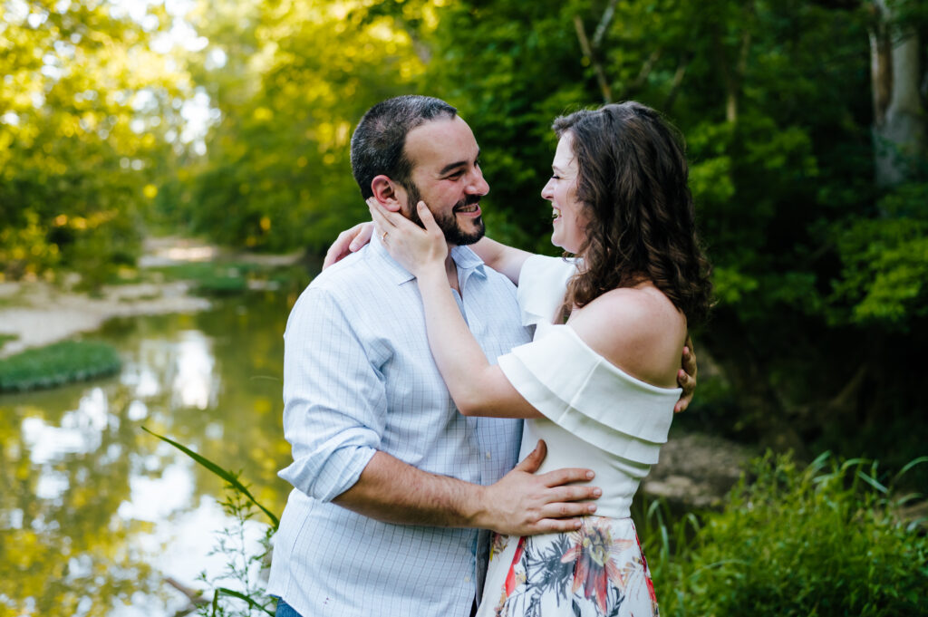 A man and woman embrace in front of a beautiful creek surrounded by greenery.