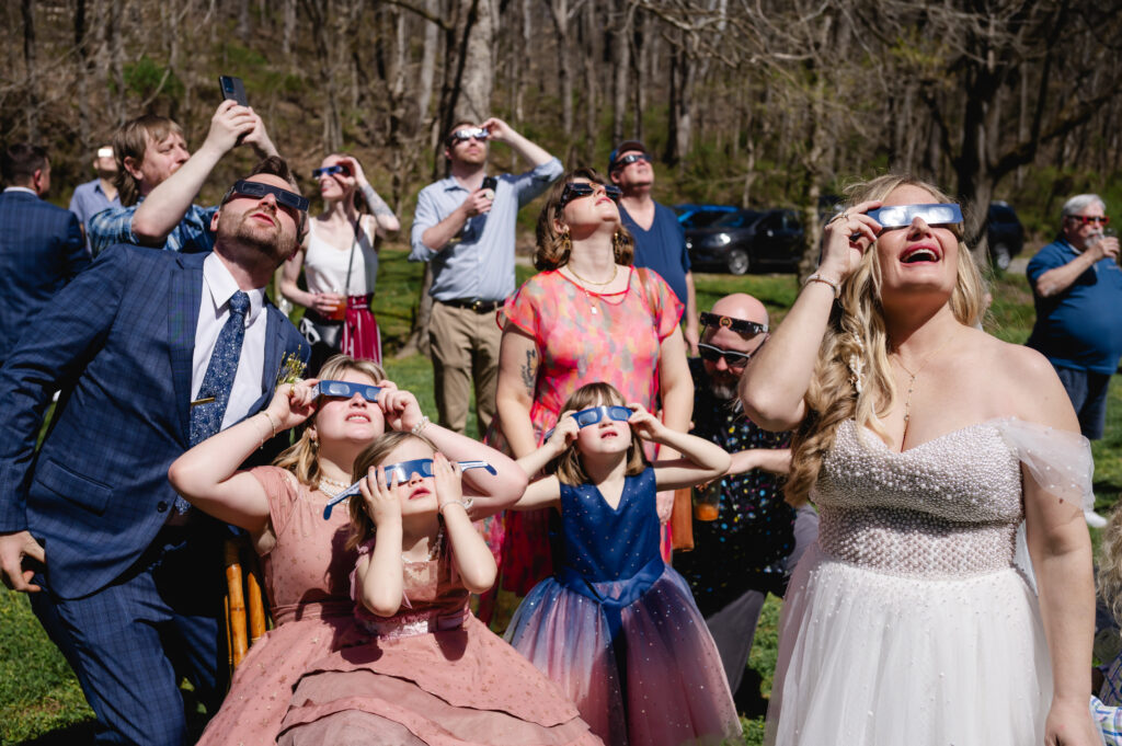 Newlyweds and their guests wear solar eclipse glasses and gaze up at the sky