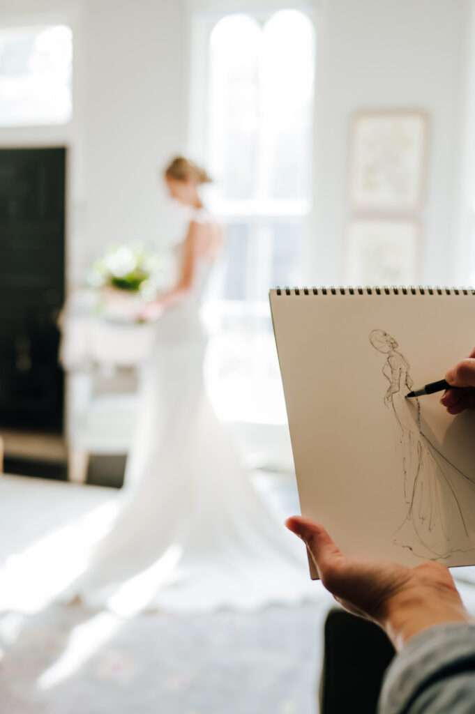 A bride poses elegantly in a bright room with tall windows, as an artist freehand sketches her in the foreground.