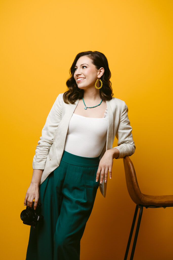 A photographer holding her camera smiles coyly into the distance. She wears a styish ivory blazer, a turquoise necklace and teal wide leg pants and stands in front of a golden yellow backdrop.