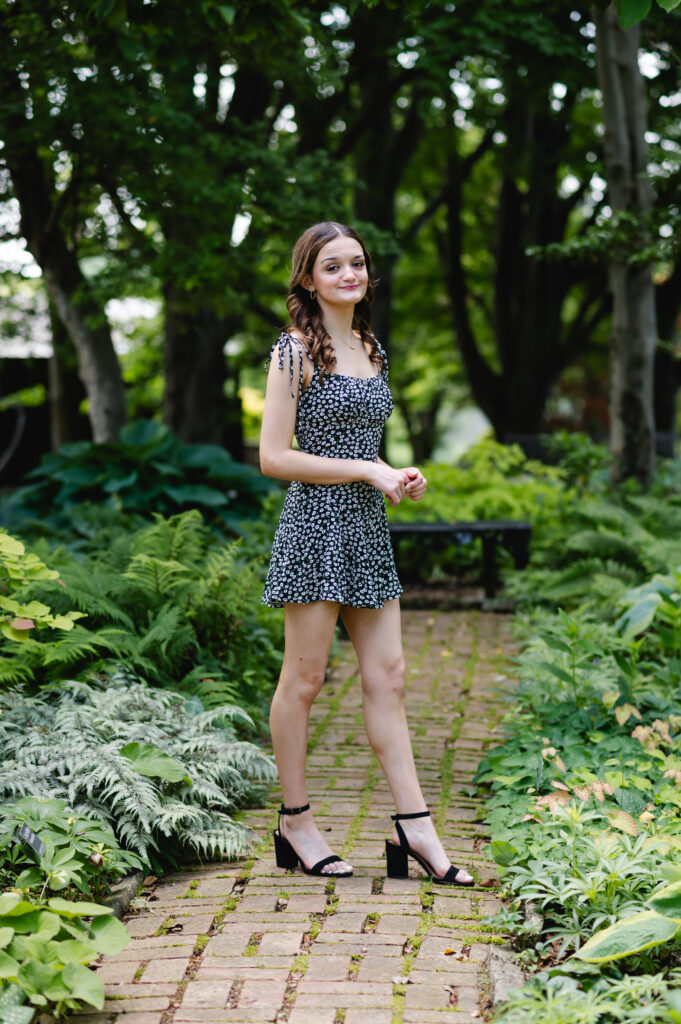 A high school senior stands among the greenery on a brick path at Yew Dell Botanical Gardens wearing her graduation cap and a floral print romper