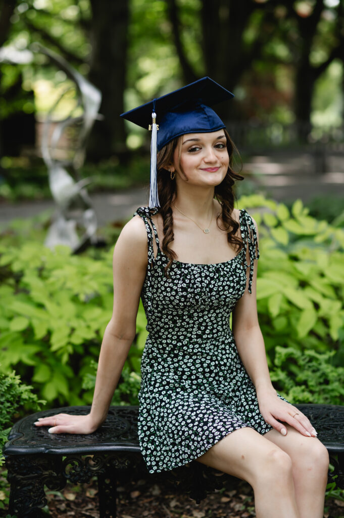 A high school senior sits on a bench at Yew Dell Botanical Gardens wearing her graduation cap and a floral print romper