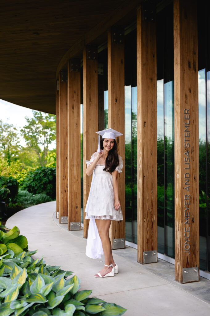 A high school grad wearing a white dress dons her cap and gown in front of the wooden and glass architectural building at Waterfront Botanical Gardens