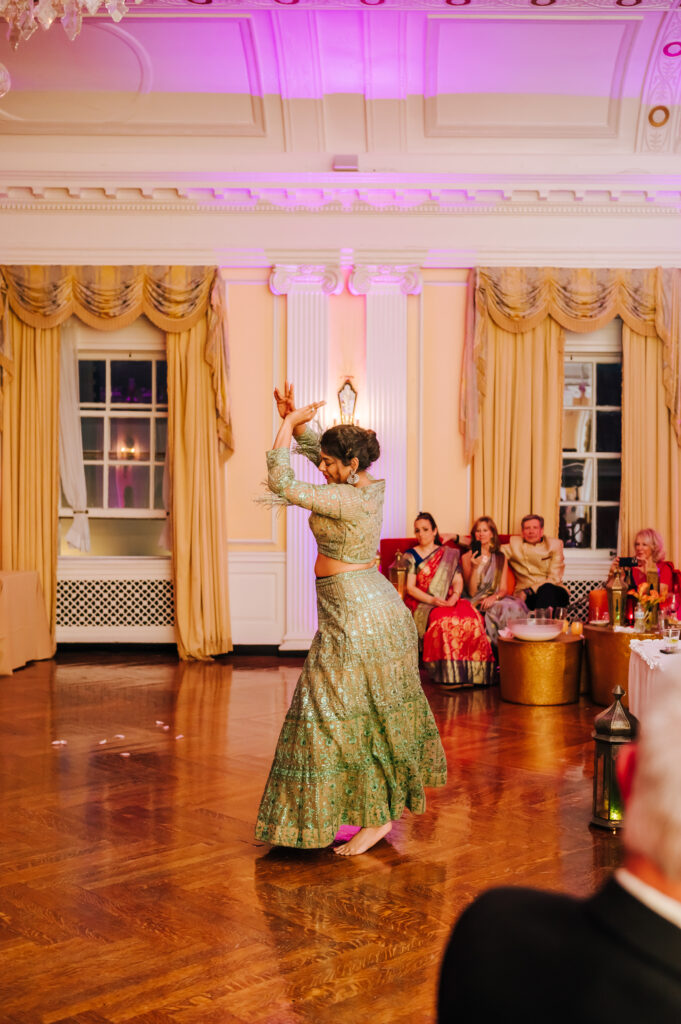 A woman wearing a green sari claps her hands in the air as she dances to a Bollywood song