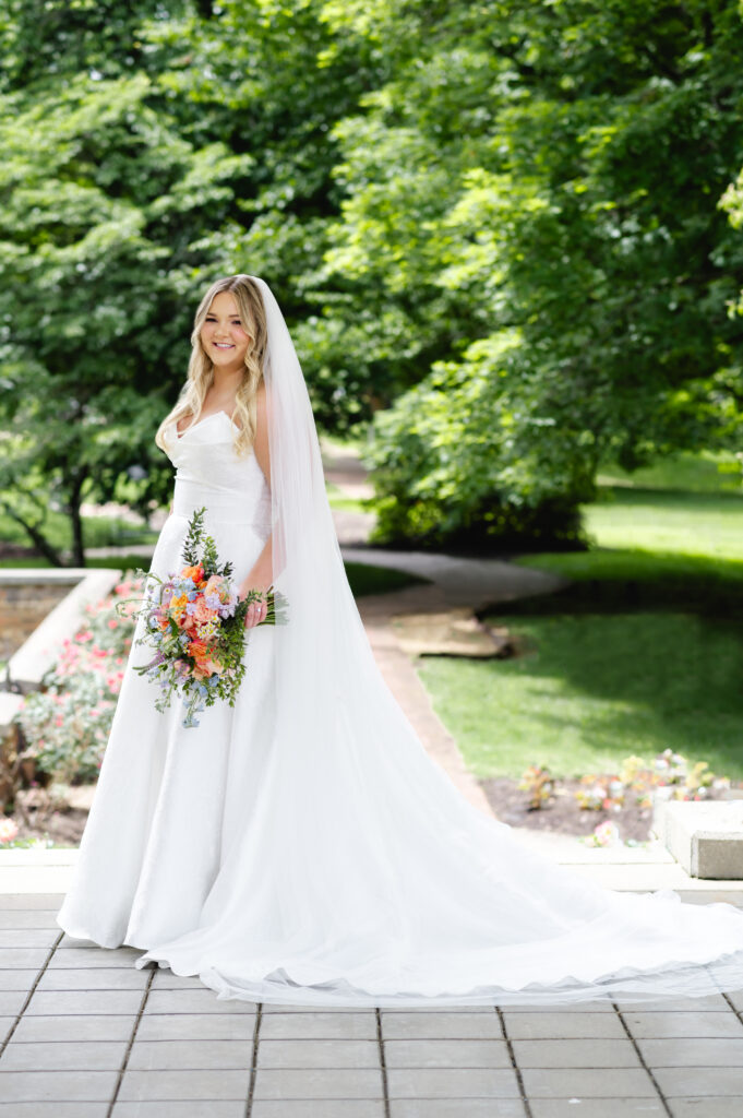 A bride stands radiantly on the veranda of My Old Kentucky Home in front of beautiful green trees