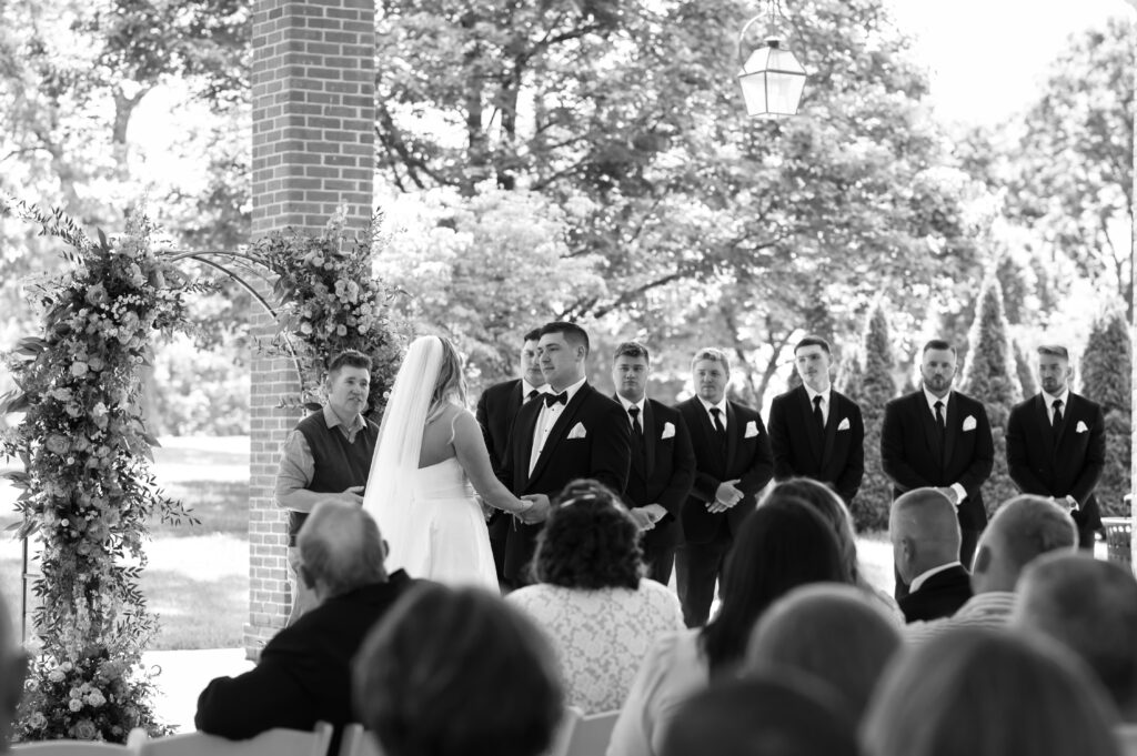 A black and white photo of the couple holding hands from the crowd's perspective as they say their vows