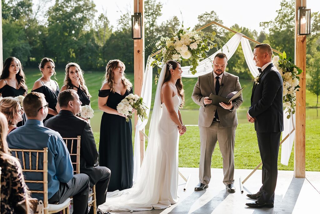 A bride and groom smile during an outdoor wedding ceremony.