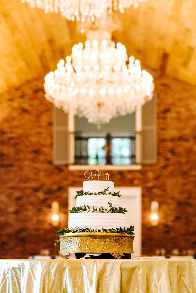 A tiered wedding cake sits on a table under a chandelier in a reception hall.