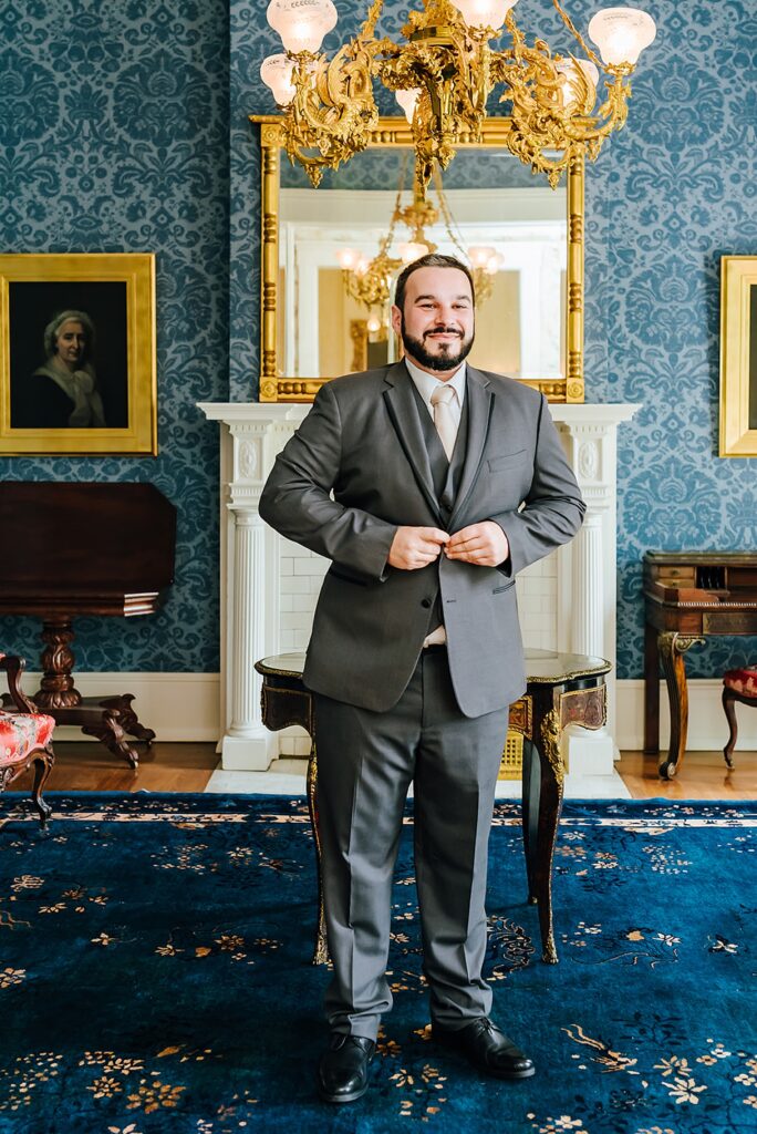 A groom buttons his jacket inside a historic wedding venue decorated with blue wallpaper in Louisville.