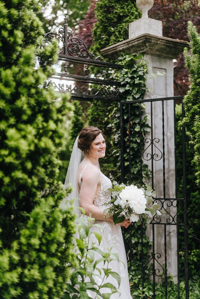 A bride stands in a garden gate smiling over her shoulder at a Louisville wedding venue.