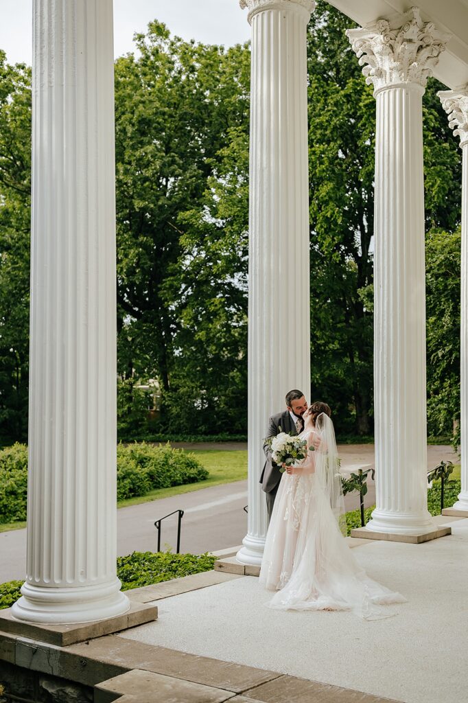 A bride and groom embrace on a porch lined by columns outside a Louisville wedding venue.