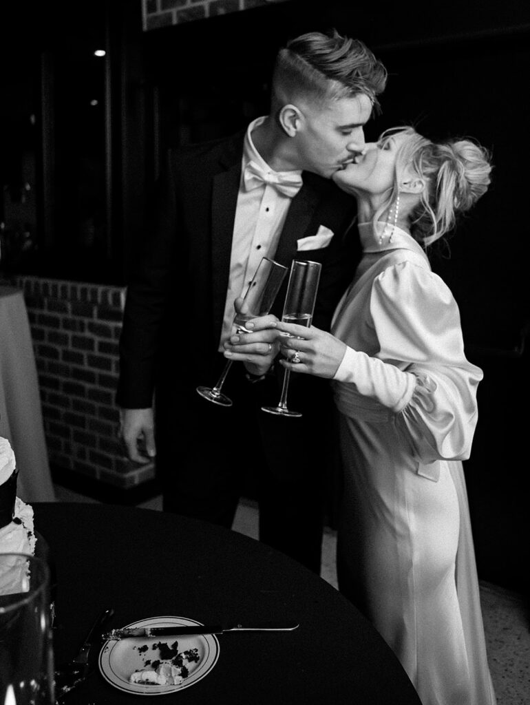 A bride and groom kiss beside a table with a slice of cake on it.