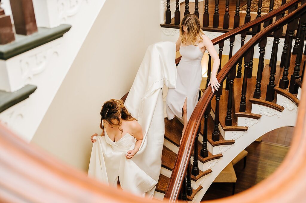 A bridesmaid carries a bride's train down a staircase.