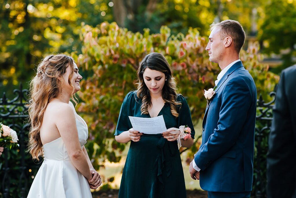 A bride and groom look at each other while an officiant reads during a wedding ceremony in a garden.