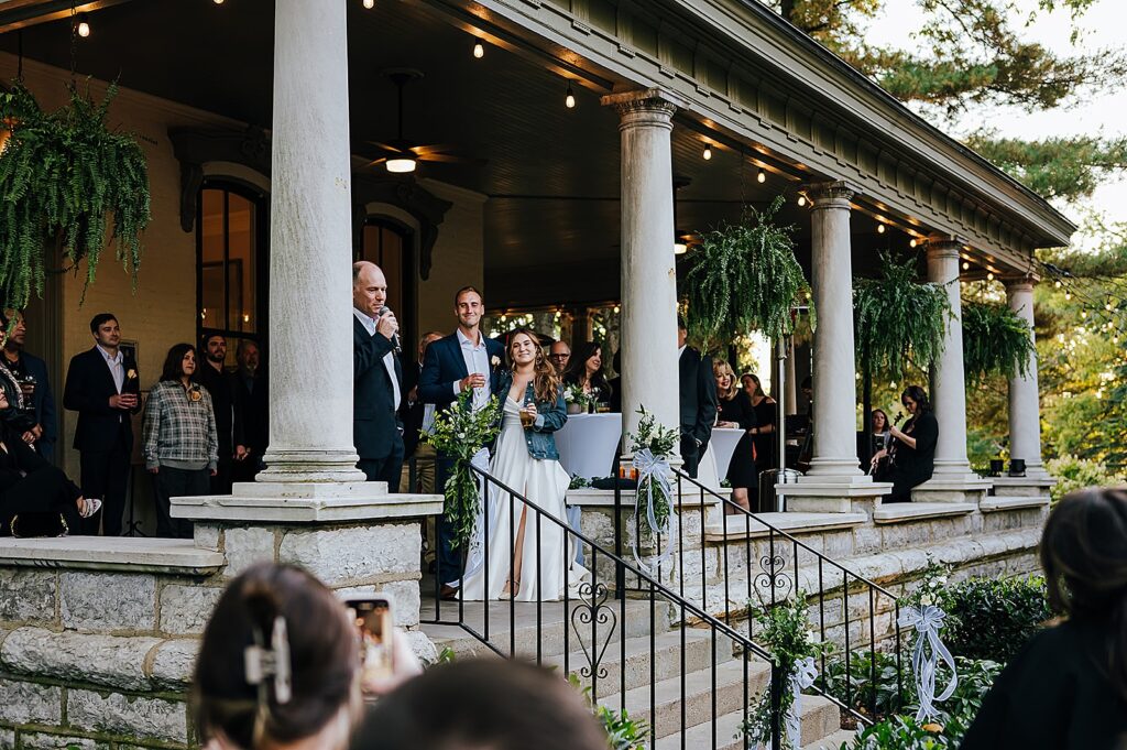 A bride and groom stand on the porch of a historic house and listen to a wedding toast.