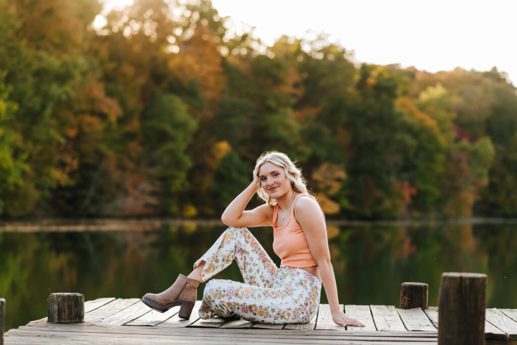 A girl in a pink top and flower patterned pants sits on the dock in front of a row of colorful fall trees