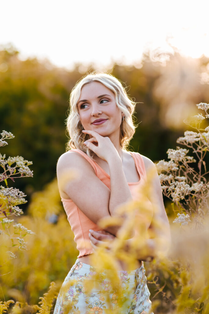 A girl with a pink top and flower patterned pants stands in a field of goldenrod and places on hand on the side of her face