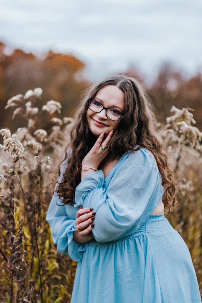 A girl with glasses wearing a light blue dress stands in a field of dried goldenrod and places on hand on the side of her face