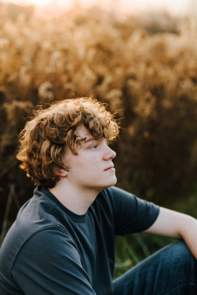 A teen boy looks to the right as he sits in a field of dried goldenrod. His golden hair matches the glow of the sun through the goldenrods behind him.