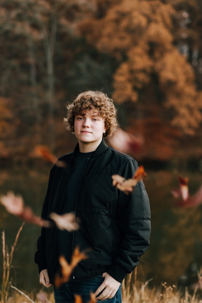 An artsy photo of a senior boy standing and looking seriously at the camera as colorful auburn leaves fall in the foreground