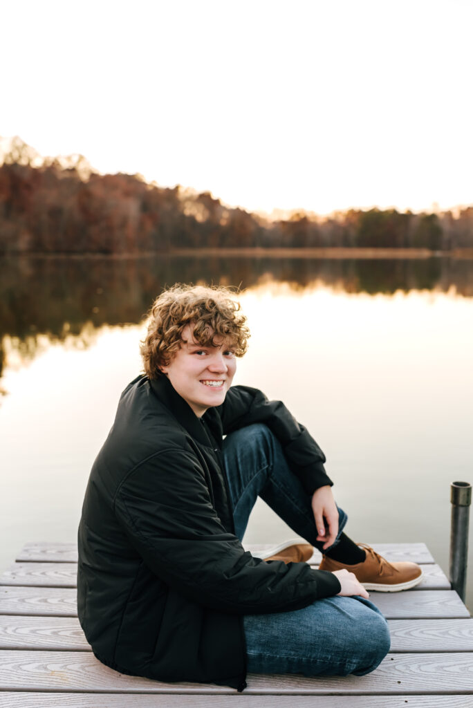 A teen boy sits on the edge of a dock overlooking a peaceful lake and smiles towards the camera