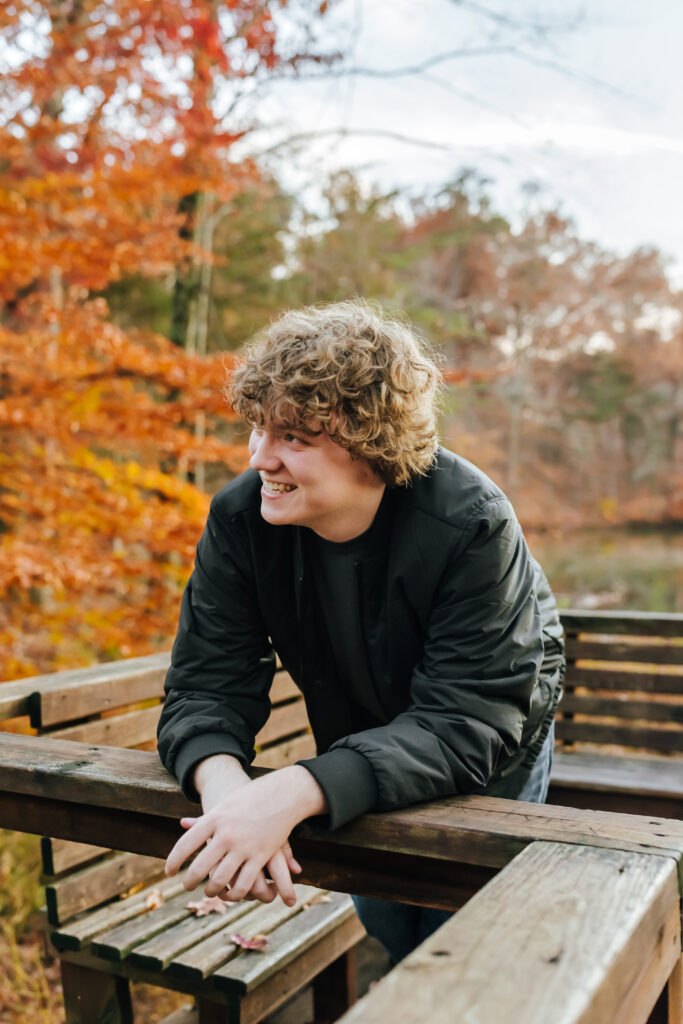 A high school senior boy wearing a black bomber jacket smiles and looks out past the camera, with glowing orange leaves behind him in the background.