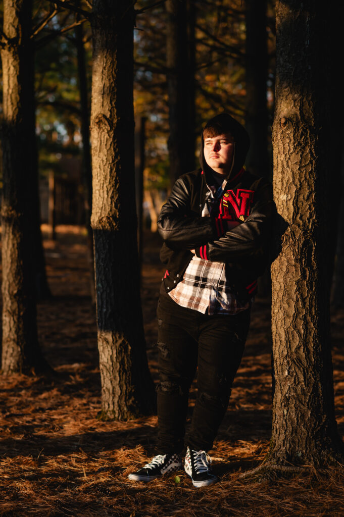 A senior boy wearing a letterman jacket leans against a pine tree as the afternoon sun illuminates his face