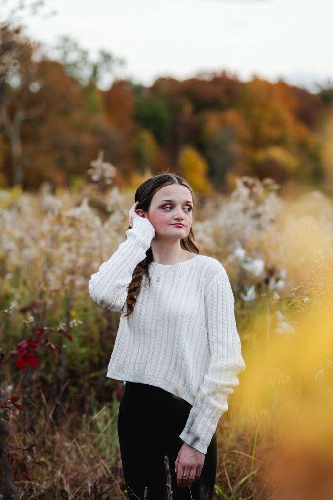 A girl in a white sweater stands in a field of dried goldenrod and places on hand on the side of her face, looking into the distance