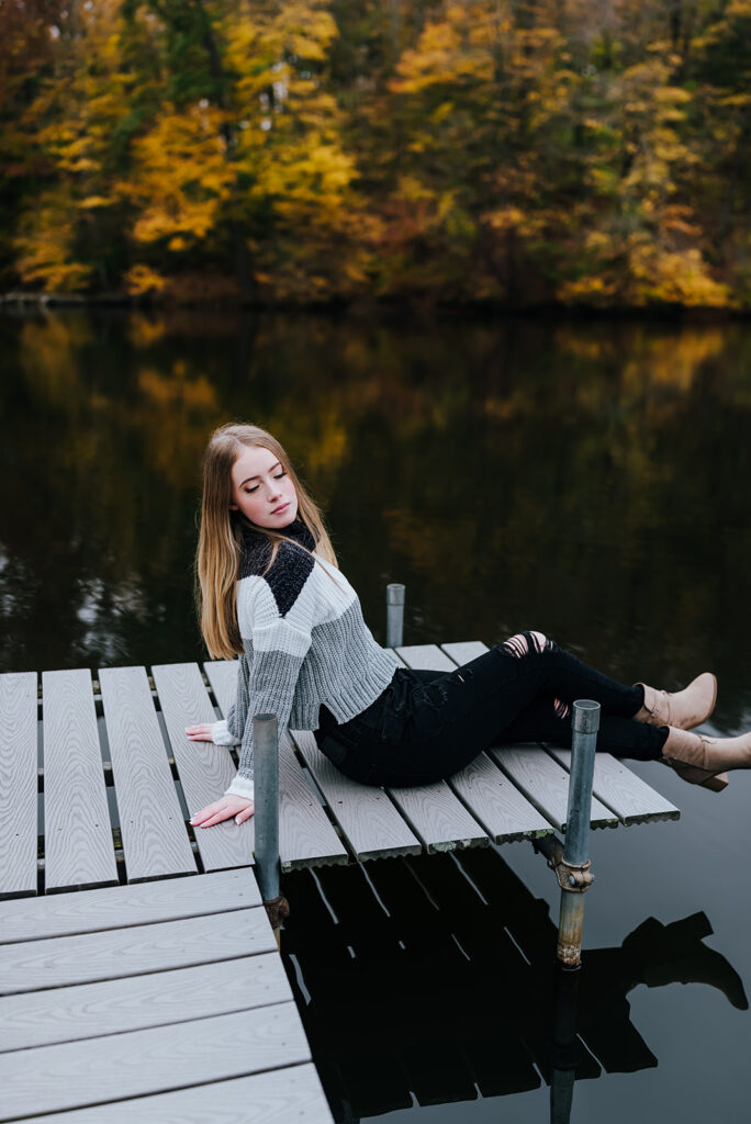 A girl in a striped sweater and black jeans looks downward as she sits on a dock in front of a still lake.