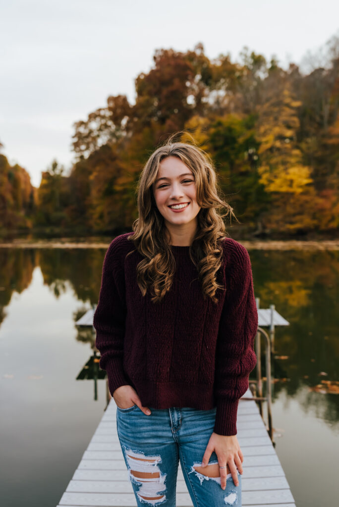 A girl in a burgundy sweater and jeans smiles with one hand in her pocket and the other by her side. She stands on a dock in front of a row of colorful fall trees.