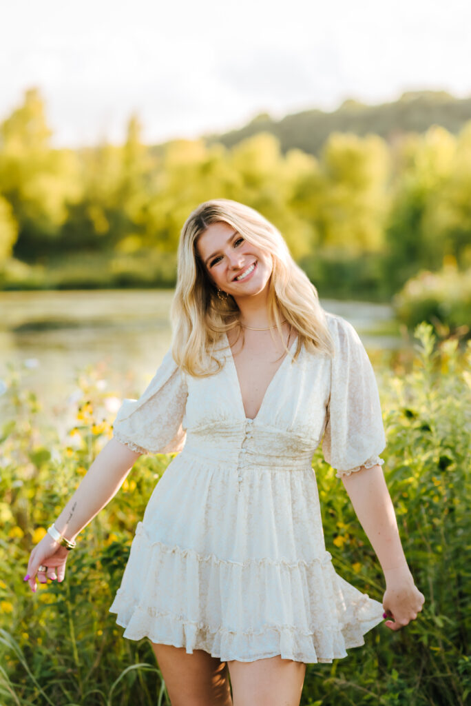 A radiant grad smiles as she frolics in a field of tall grass by a small bond.
