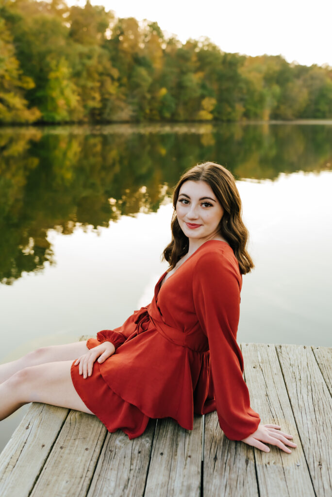 A girl in a burnt orange dress smiles softly on a dock in front of a row of green trees