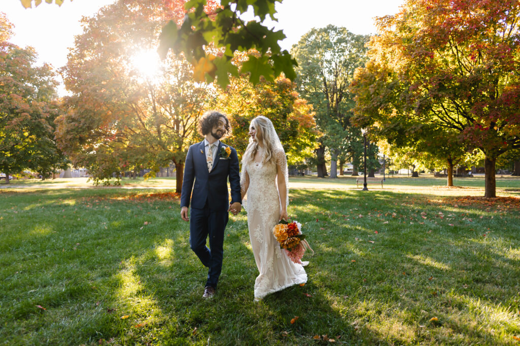 A groom in a navy suit and bride walk hand in hand through the colorful trees of Shelby Park in the fall