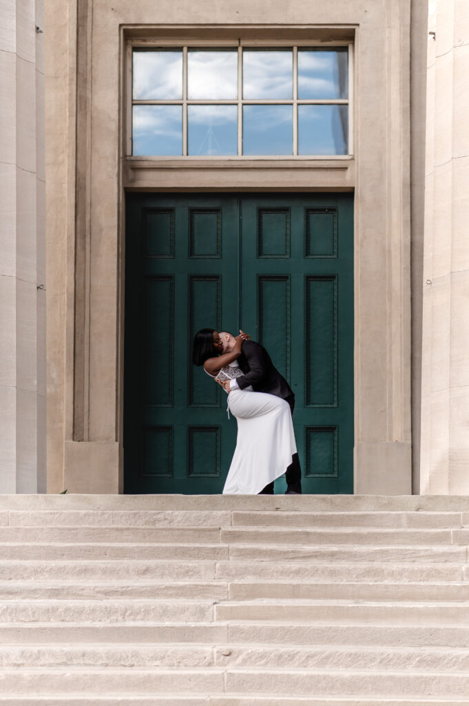 A couple dips for a kiss as they stand atop the stairs of Metro Hall in front of a giant teal door