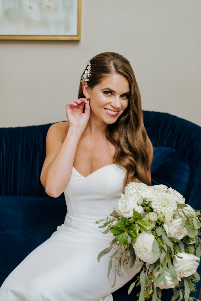 A bride adjusts her earring as she holds her flowers. She sits on an elegant velvet blue couch.