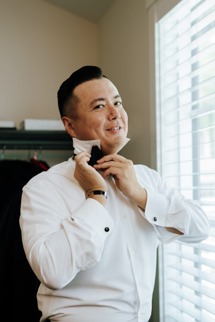 A groom looks at the camera as he puts on his bowtie 